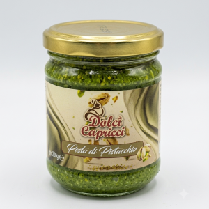 Un barattolo di vetro contenente pesto di pistacchio, con un tappo di metallo dorato, fotografato in primo piano su uno sfondo bianco nitido. L'etichetta beige sul barattolo mostra il logo "Dolci Capricci" in lettere rosse corsive ed eleganti, sopra la scritta "Pesto di Pistacchio" in bianco all'interno di un cartiglio marrone. Sull'etichetta sono illustrati dei pistacchi interi e in granella. Il pesto all'interno del barattolo ha una consistenza densa e un colore verde brillante e intenso. In basso a sinistra sull'etichetta è visibile la dicitura "200g e".